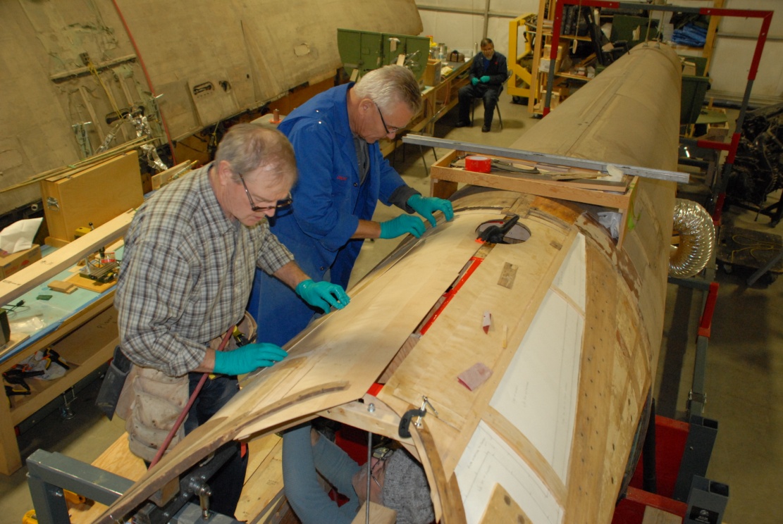 Roger, Dick and Gary (inside the fuselage) fit and install the new inner skin to the right side of the upper fuselage, aft of the cockpit area.  In the background David continues the work of stripping and restoring hydraulic components.